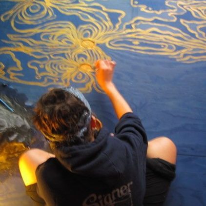 Shot from above of a dark-haired person in a sweatshirt sitting cross-legged on a blue floor drawing squiggly gold lines on the floor.
