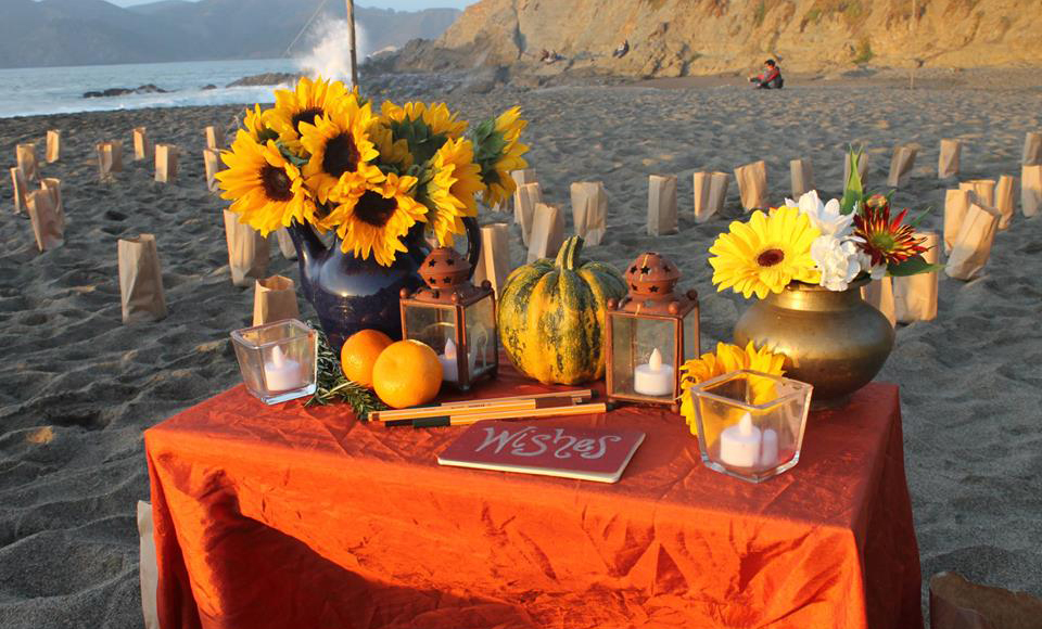A table draped with an orange cloth, on which stands several bunches of flowers and some small notebooks and pens. In the background is a sandy beach and a glimpse of waves and sandstone cliffs.
