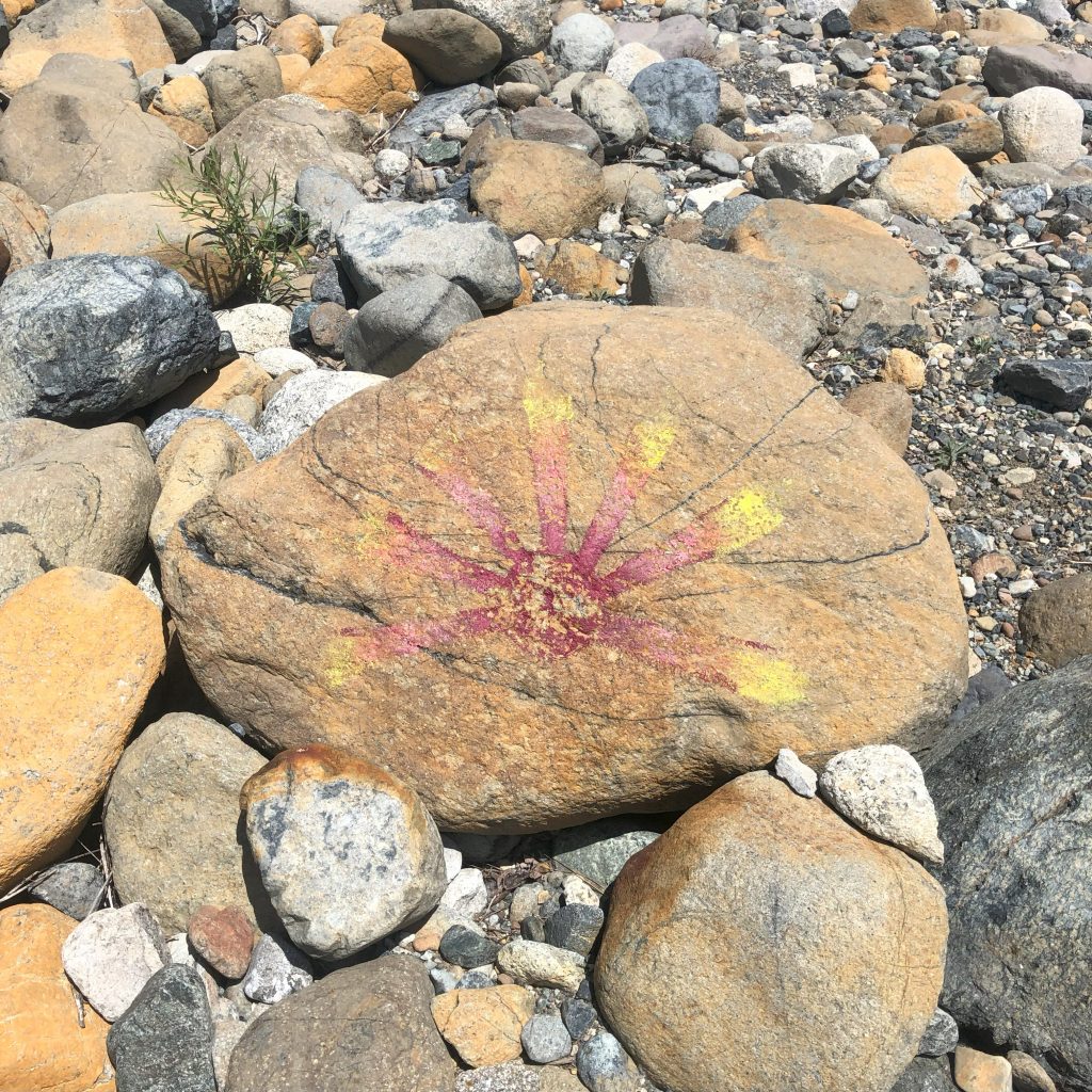 a closeup of a large orange rock with a slightly faded red and yellow sunburst painted on it. Other rocks sit around it.